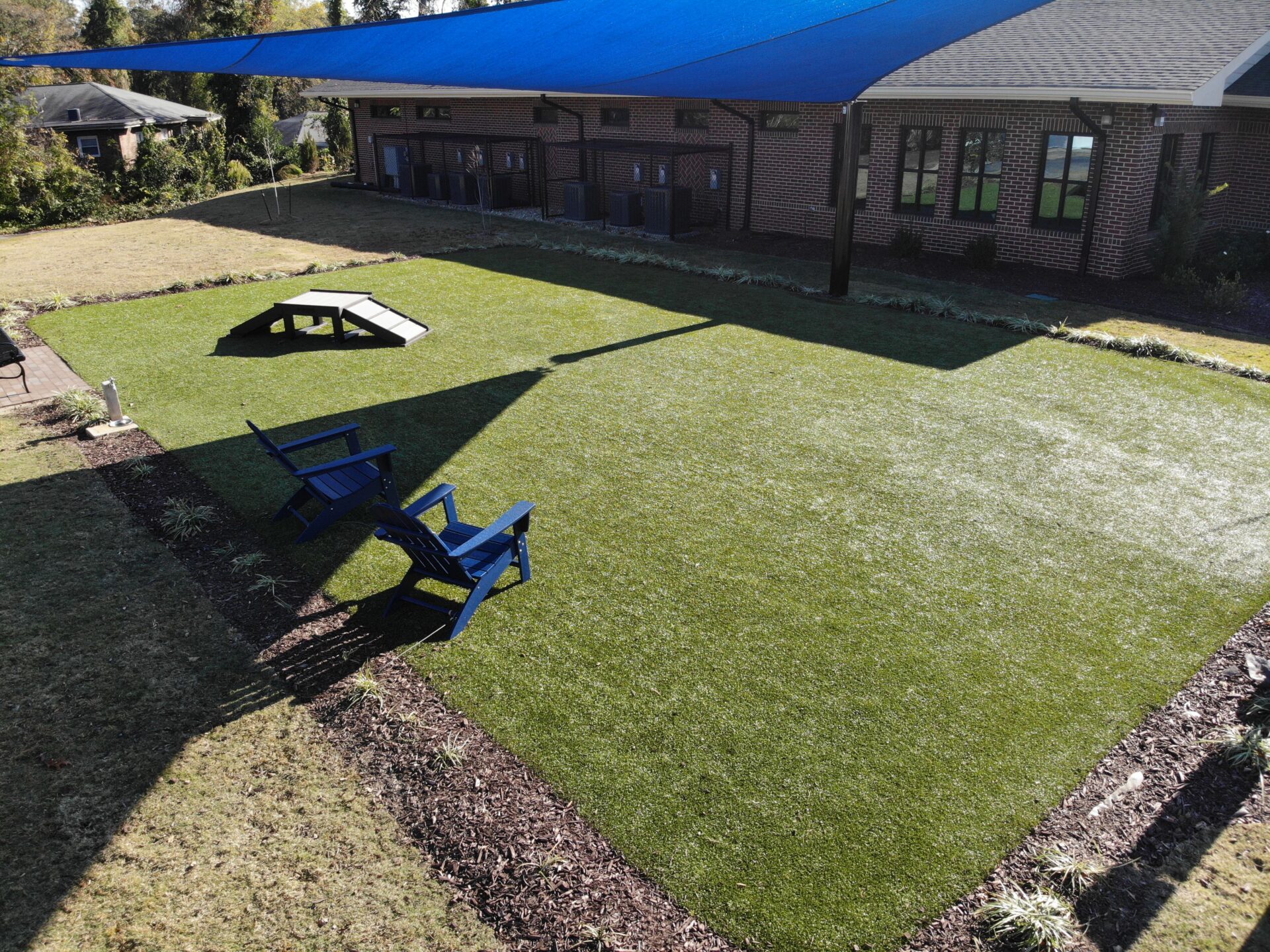 Spacious artificial turf play yard with shade sails, outdoor seating, and a small agility ramp beside a brick building.