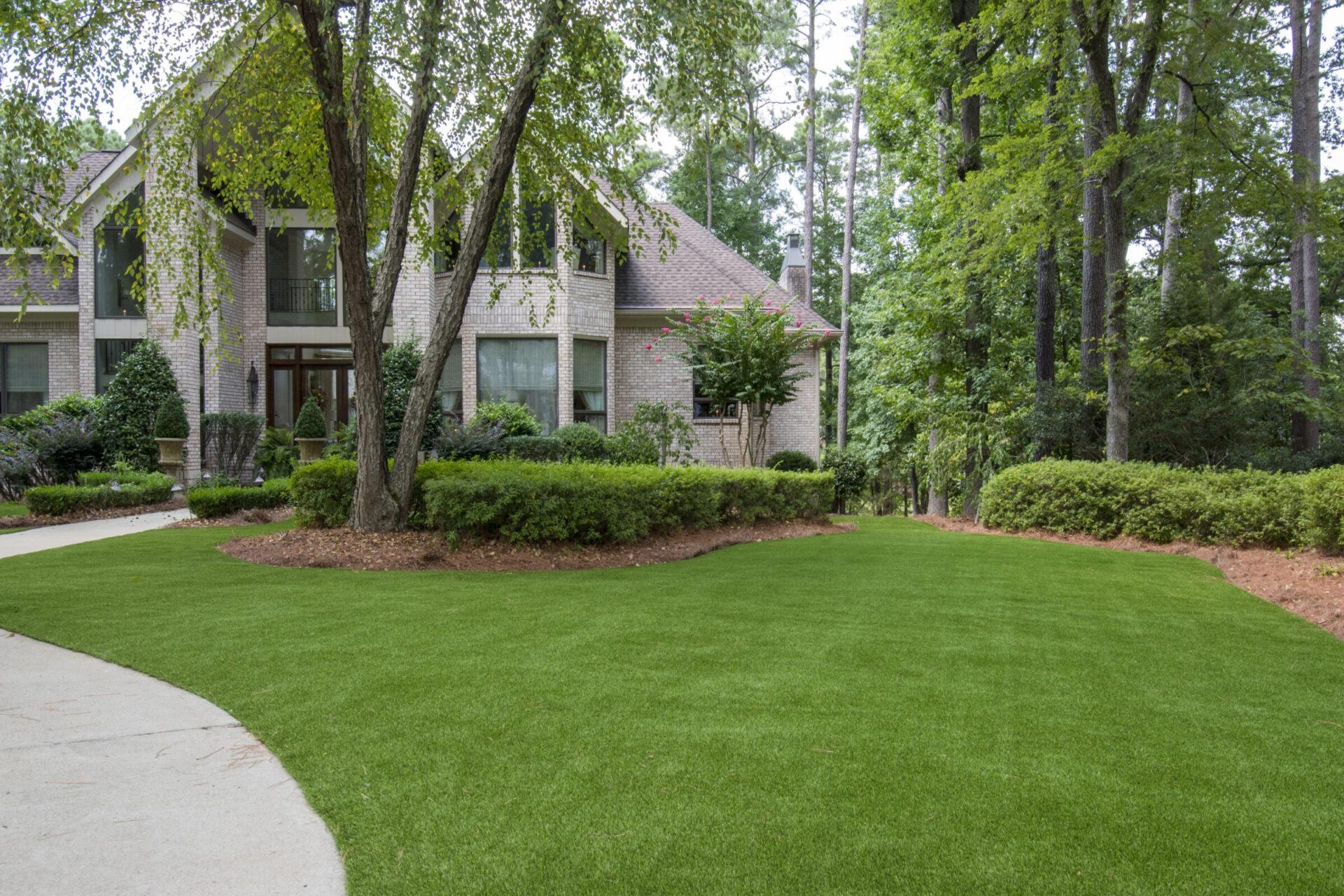 Artificial turf front lawn surrounding a large brick home with mature trees, shrubs, and a curved sidewalk.