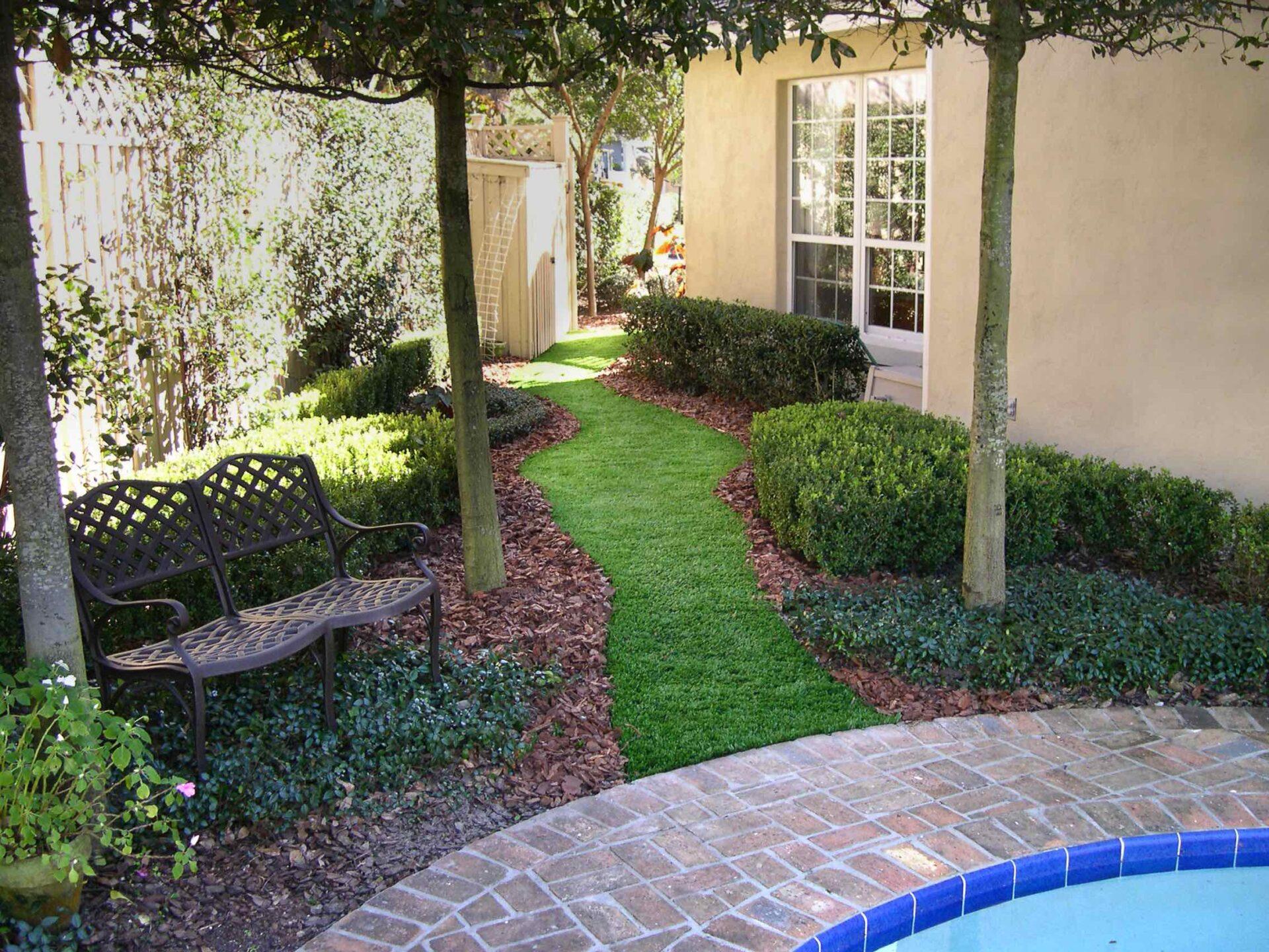 Curved artificial turf pathway bordered by hedges, trees, mulch, and a brick patio area with a garden bench.